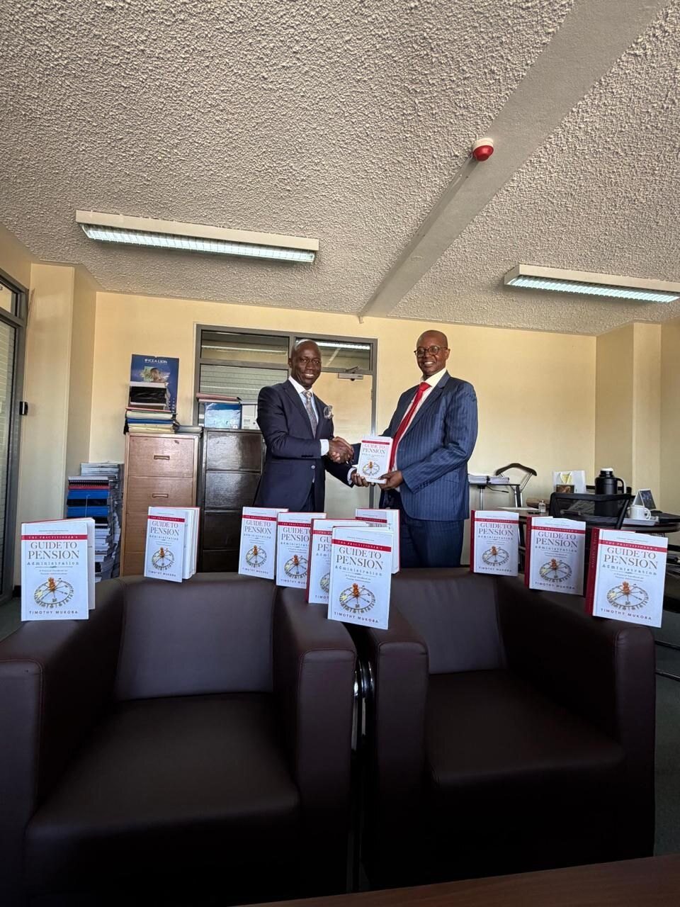 Ten copies of The Practitioner’s Guide to Pension Administration lined up in an office, with two men shaking hands behind them.