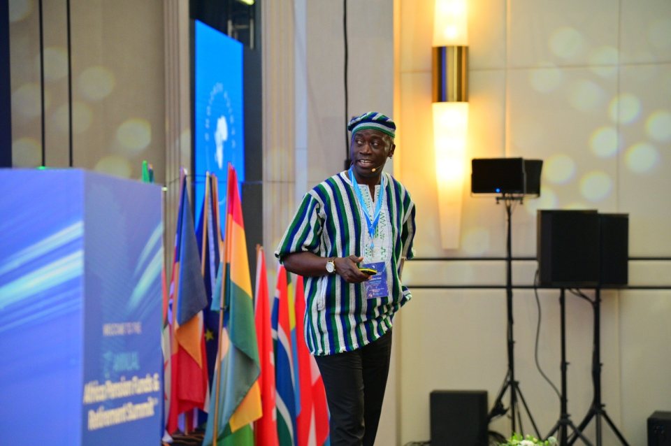 Timothy Mukoba, in a striped smock and cap, presenting beside a row of national flags at the summit.